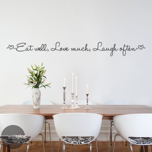 May include: A white wall with the text "~Eat well, Love much, Laugh often~" in black script with a heart symbol at the beginning and end of the text. A wooden dining table with white chairs and a vase of white flowers sits in front of the wall.