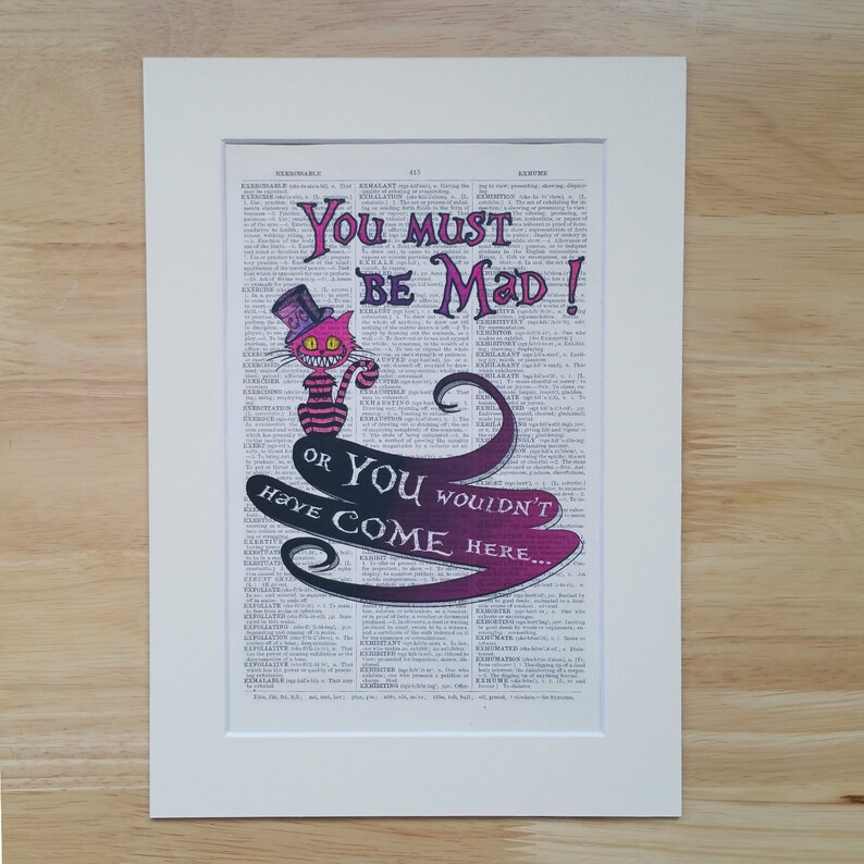 Pu&ograve; includere: Una pagina di un dizionario vintage con un'illustrazione del Gatto del Cheshire. Il gatto indossa un cappello a cilindro e ha un sorriso malizioso. Il testo recita "You must be mad! Or you wouldn't have come here..."