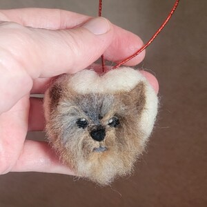 Peut inclure: Une décoration en forme de cœur en feutre avec le visage d'un chien, avec une fourrure brune, blanche et grise. La décoration a une ficelle rouge pour l'accrocher.
