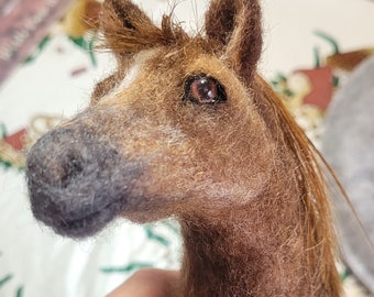May include: Close-up of a brown and white needle felted horse with a dark gray nose and brown eyes. The horse has a long brown mane and tail.