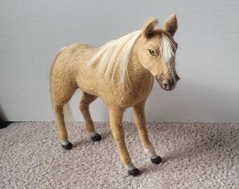 May include: A hand-felted horse figurine with a light brown body and a white mane and tail. The horse has black hooves and is standing on a beige carpet.