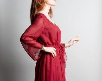 May include: A woman wearing a long, flowing red dress with a cinched waist and long sleeves. The dress has a beaded trim on the sleeves and a flowing skirt.
