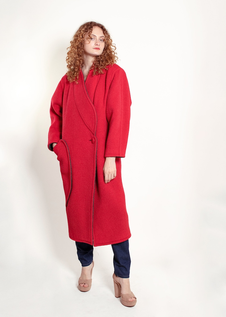 May include: A long red wool coat with a contrasting dark trim. The coat has a single button closure and a large pocket on the left side.