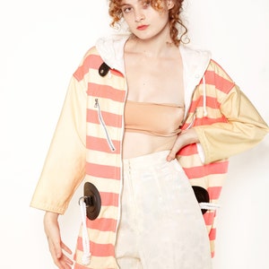 K&ouml;nnte beinhalten: Eine rosa-gelb gestreifte Jacke mit einer wei&szlig;en Kapuze und schwarzen Akzenten. Die Jacke hat einen Rei&szlig;verschluss und zwei gro&szlig;e schwarze Kreise auf der Vorderseite. Das Model tr&auml;gt eine wei&szlig;e Hose mit Blumenmuster.