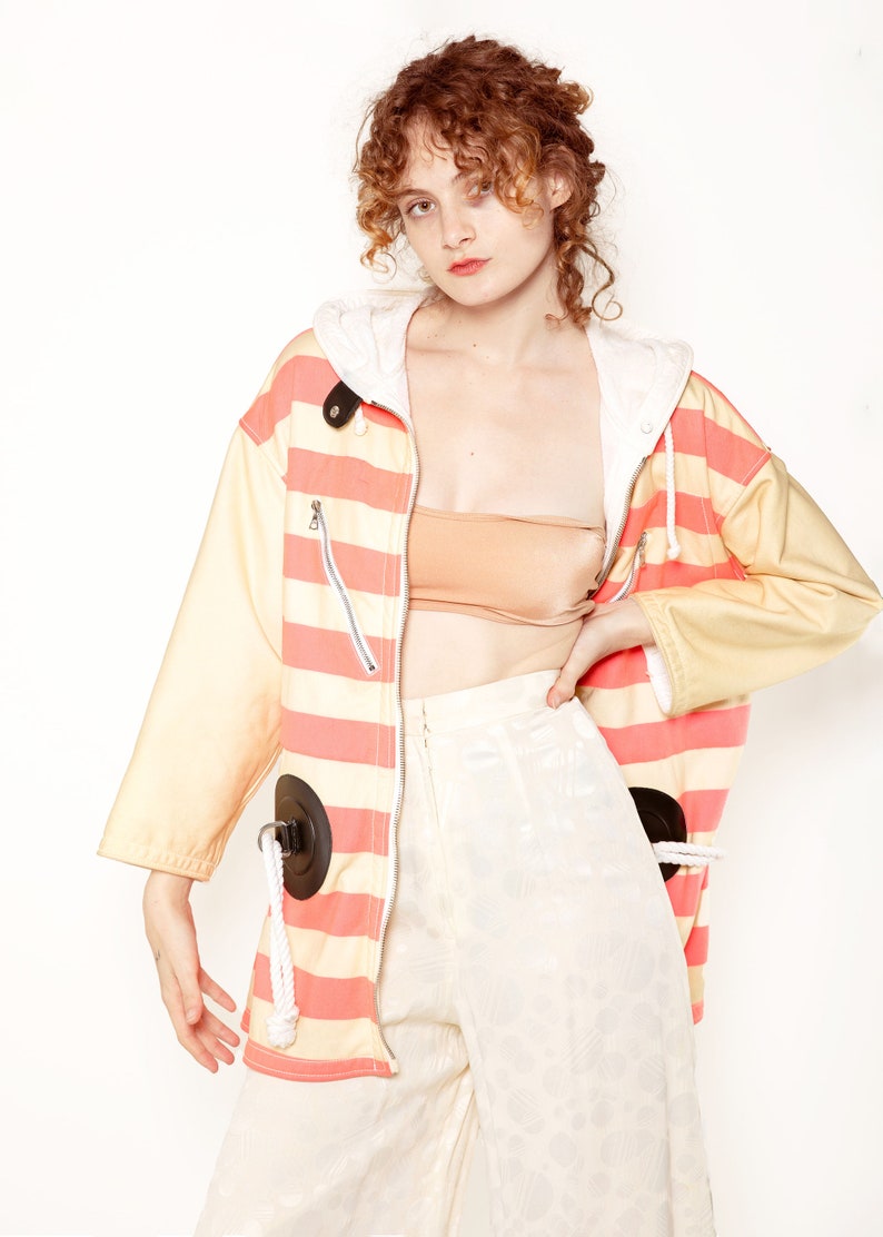 K&ouml;nnte beinhalten: Eine rosa-gelb gestreifte Jacke mit einer wei&szlig;en Kapuze und schwarzen Akzenten. Die Jacke hat einen Rei&szlig;verschluss und zwei gro&szlig;e schwarze Kreise auf der Vorderseite. Das Model tr&auml;gt eine wei&szlig;e Hose mit Blumenmuster.