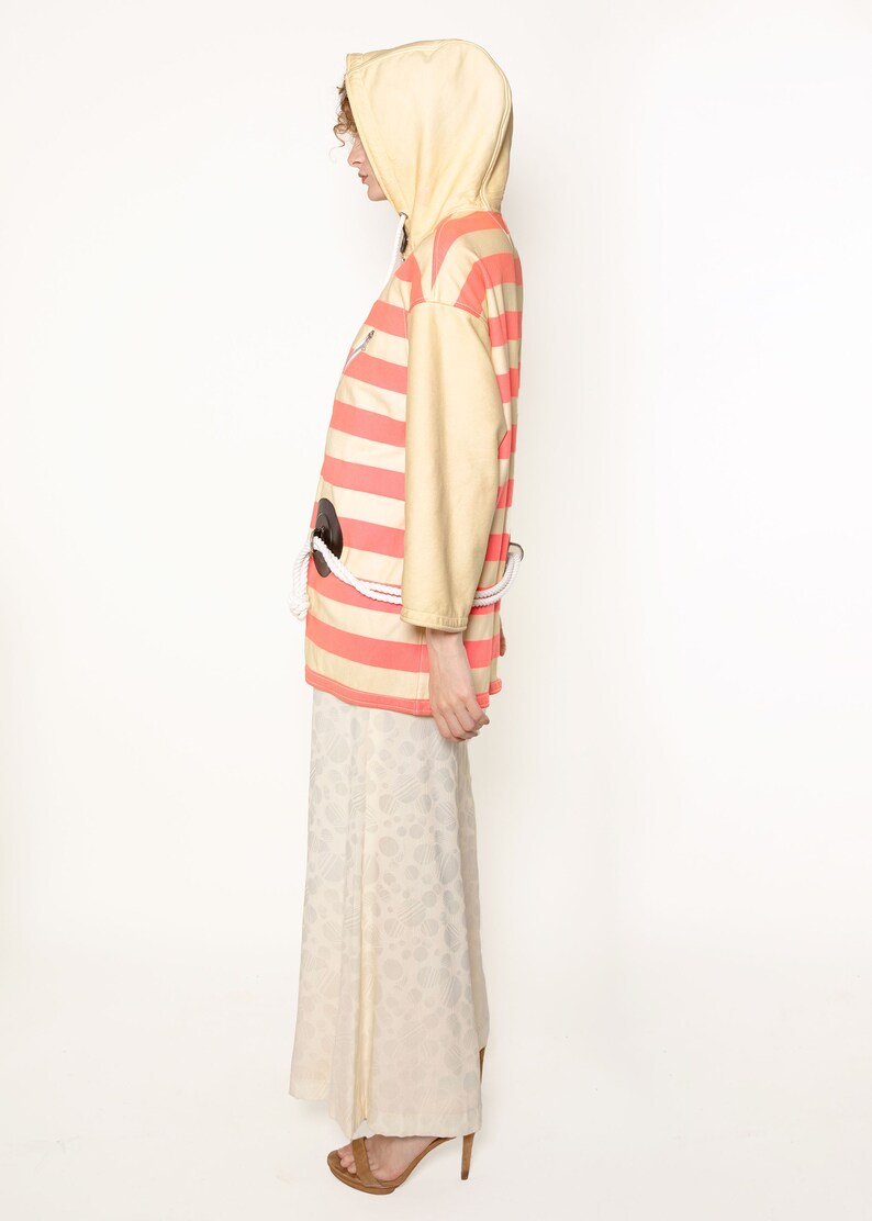 K&ouml;nnte beinhalten: Eine gestreifte Kapuzenjacke mit einem Seilg&uuml;rtel. Die Jacke ist gelb und rosa mit horizontalen Streifen. Das Model tr&auml;gt einen wei&szlig;en Rock mit Blumenmuster.