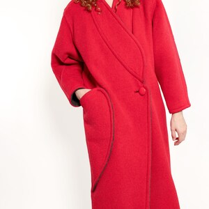 May include: A long red wool coat with a contrasting black trim and a single button closure. The coat has a unique asymmetrical design with a curved pocket on the left side.