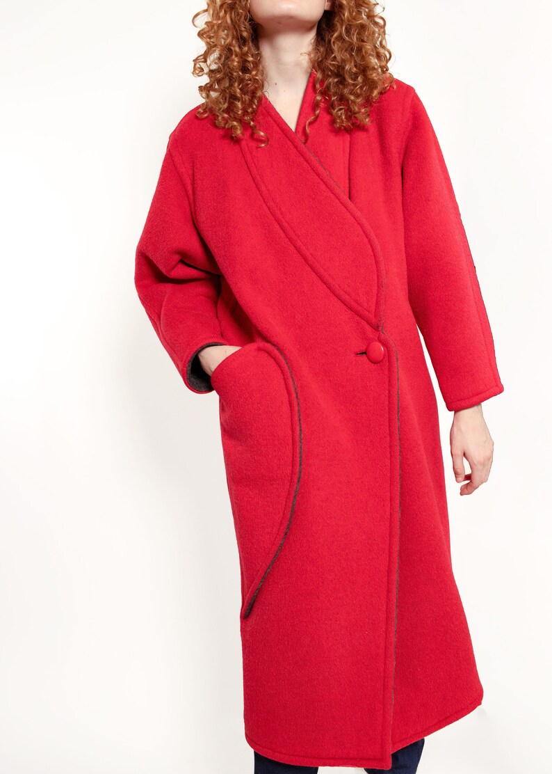 May include: A long red wool coat with a contrasting black trim and a single button closure. The coat has a unique asymmetrical design with a curved pocket on the left side.