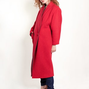 May include: A woman wearing a long, red coat with a dark gray lining. The coat has a collar and a single button closure. She is wearing blue jeans and brown heeled sandals.