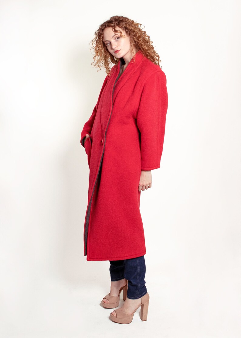 May include: A woman wearing a long, red coat with a dark gray lining. The coat has a collar and a single button closure. She is wearing blue jeans and brown heeled sandals.