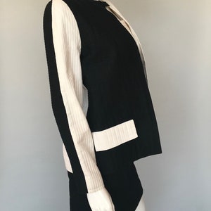 May include: A black and white skirt suit with a white stripe down the sleeves. The jacket has a round neckline and a single button closure.