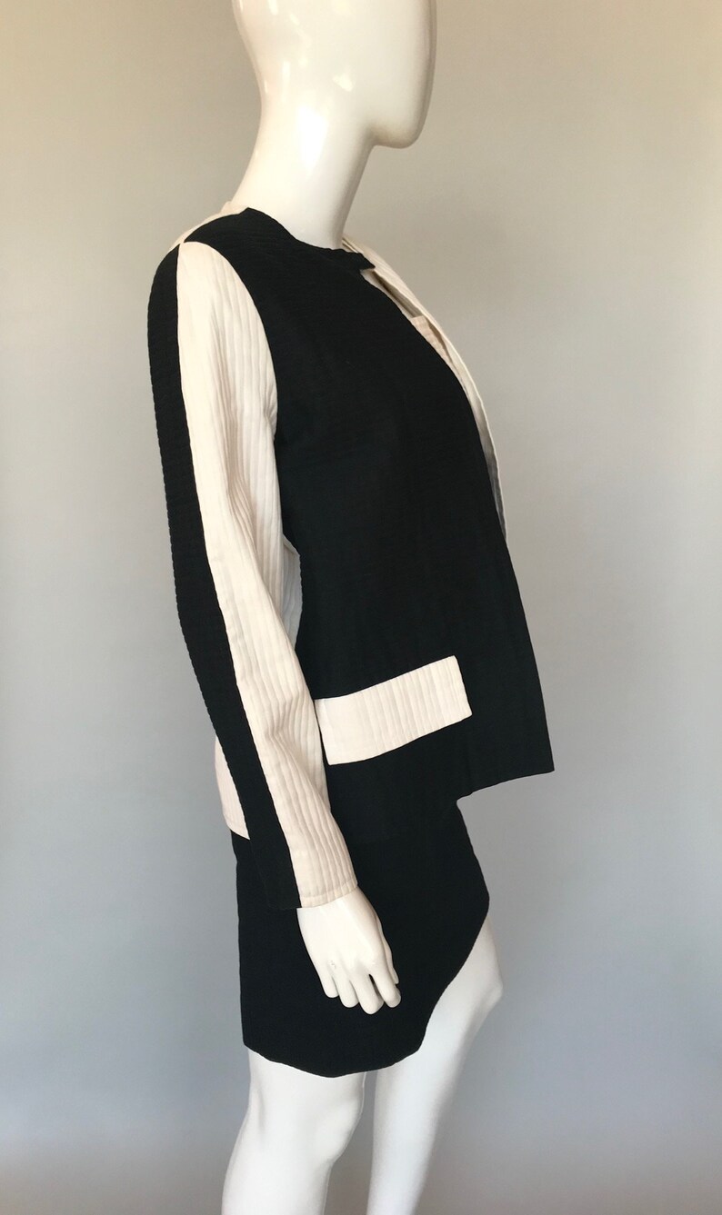 May include: A black and white skirt suit with a white stripe down the sleeves. The jacket has a round neckline and a single button closure.