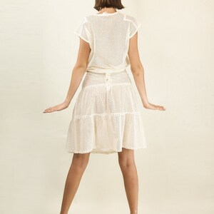 May include: A white crochet dress with a tiered skirt and a fitted bodice. The dress is made of a lightweight, breathable fabric and has a relaxed fit. The model is wearing the dress with a pair of teal platform heels.