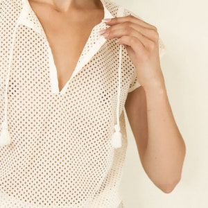 May include: A white mesh top with a V-neck and short sleeves. The top has a drawstring at the waist and tassels at the neckline.
