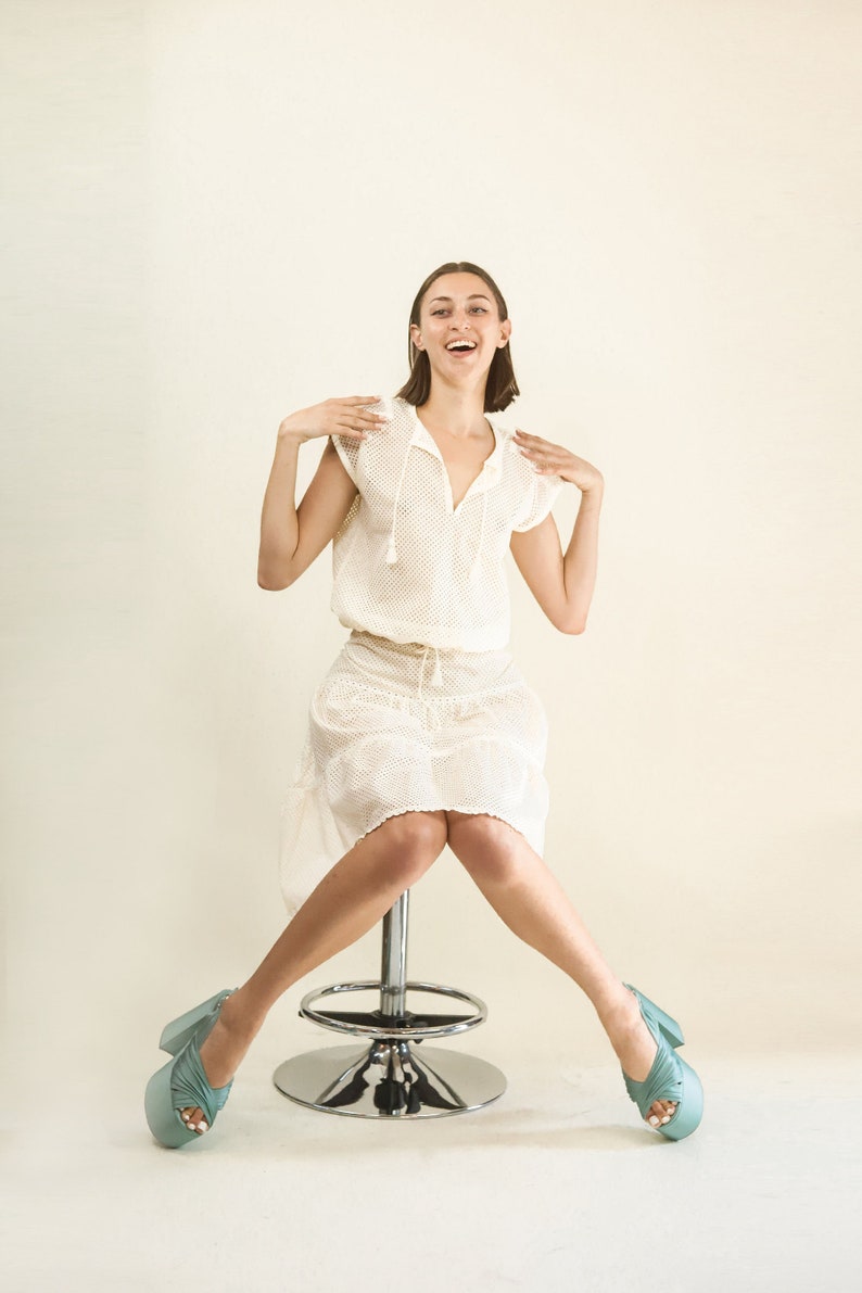 May include: A woman wearing a white crochet dress and light blue platform shoes sits on a silver stool with her legs crossed.