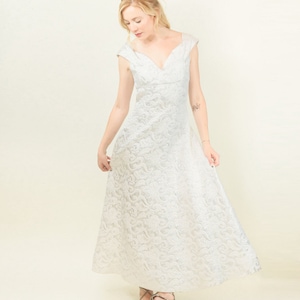 Puede incluir: Una mujer con un vestido blanco con estampado plateado, cuello en V y mangas cortas. Lleva tacones dorados y está de pie sobre un fondo blanco.
