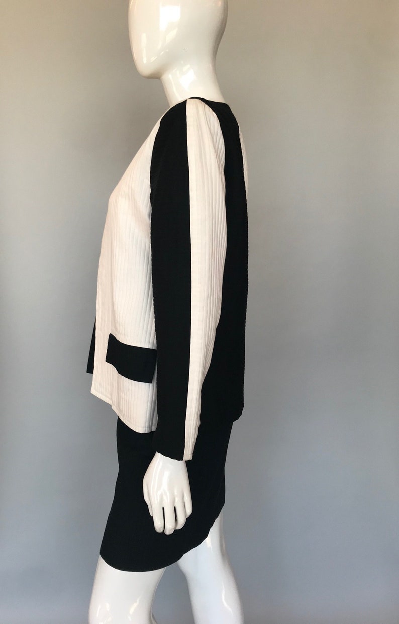 May include: A white and black skirt suit with a ribbed texture. The jacket has a black panel down the side of each sleeve. The skirt is a simple A-line style.