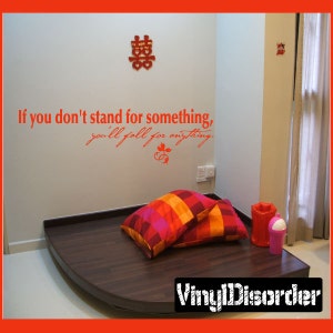 May include: A red and orange striped pillow and a red pillow with a white and pink checkered pattern are on a dark brown wooden platform. The wall behind the platform is white with a red Chinese character and the text "If you don't stand for something, you'll fall for anything." in black.