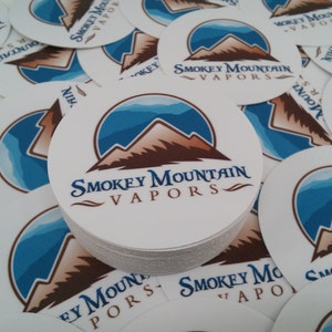 May include: A stack of round white stickers with a blue and brown mountain design. The stickers feature the text "Smokey Mountain Vapors" in a stylized font.