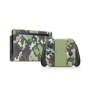 Op de afbeelding: Een gaming console en controllers met een camouflagepatroon. De console en controllers hebben een groen, bruin en grijs camouflageontwerp. De controllers hebben zwarte handgrepen en knoppen. De console staat op een witte achtergrond.