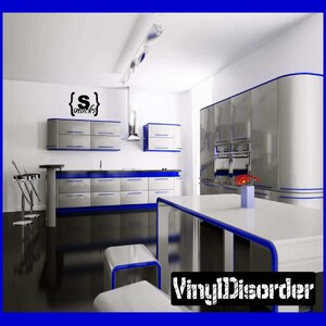 May include: A modern kitchen with white cabinets and blue trim. The cabinets are open and reveal a stainless steel refrigerator and oven. There is a table in the centre of the room with a vase of flowers on it. The wall has a black vinyl decal that says "{Snacks}".