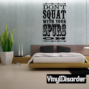 Può includere: Adesivo da parete in bianco e nero con il testo "Don't Squat With Your Spurs On" in un font in stile western. L'adesivo è su una parete blu chiaro sopra una struttura del letto in legno con biancheria da letto bianca.