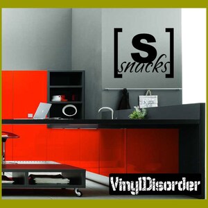May include: A black and white vinyl decal with the text "[S] snacks" on a grey wall in a kitchen. The kitchen has red cabinets and a black worktop.