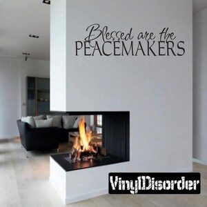 May include: A black and white vinyl wall decal with the text "Blessed are the Peacemakers" on a white wall above a modern fireplace with a fire burning inside.
