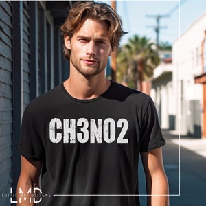 May include: A man wearing a black t-shirt with the text "CH3NO2" printed in white. The man is standing in a city street.