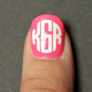May include: A pink fingernail with a white monogram sticker that says "KSR" on it.