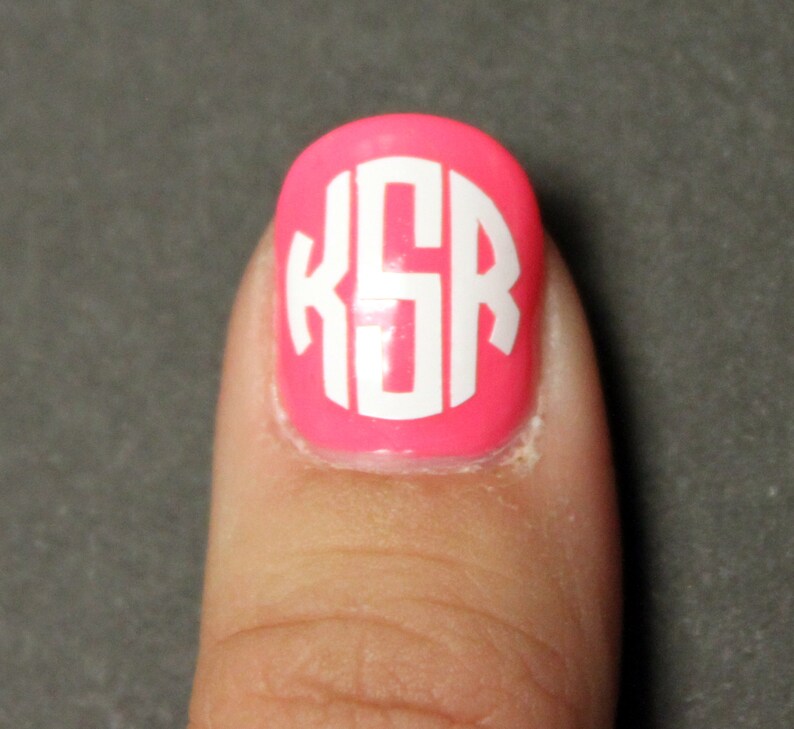 May include: A pink fingernail with a white monogram sticker that says "KSR" on it.