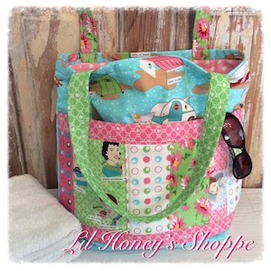 May include: A green and pink patchwork tote bag with a blue and white camping themed fabric lining. The bag has two large pockets on the front and two handles. The bag is sitting on a wooden surface with a white towel next to it.