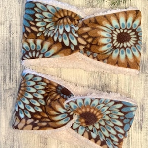 May include: Two brown and blue fleece ear warmers with a white sherpa lining. The ear warmers have a floral pattern with brown, blue, and white colors.