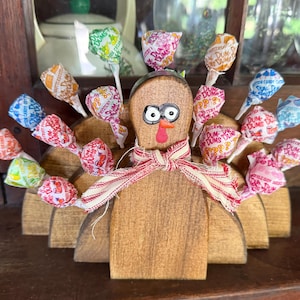 May include: Wooden turkey figurine with a brown body and a red beak. The turkey has a white and red scarf around its neck. The turkey's tail feathers are made of colorful lollipop wrappers with the text "Dum Dums" on them.