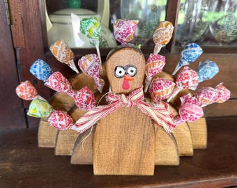 May include: Wooden turkey figurine with a brown body and a red beak. The turkey has a white and red scarf around its neck. The turkey's tail feathers are made of colorful lollipop wrappers with the text "Dum Dums" on them.