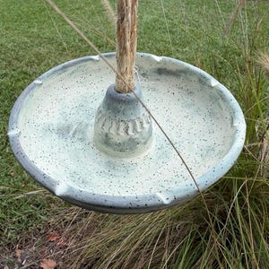 May include: A round, speckled off-white ceramic ashtray with a central conical pillar featuring vertical indentations. A thick, natural-colored jute rope extends upward from the center of the pillar, allowing the piece to be suspended.