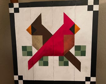 May include: A wooden barn quilt with a geometric design of two red cardinals with brown and orange accents. The cardinals are facing each other and are surrounded by green squares.