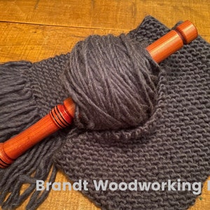 May include: A wooden yarn swift with red and black accents, wrapped with a ball of dark gray yarn and a knitted scarf.