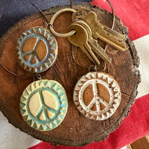 May include: Three circular ceramic pendants featuring a peace sign symbol, each with a unique glazed border and speckled clay center. The pendants are attached to a metal keyring holding two brass keys, resting on a round, textured wood slice.