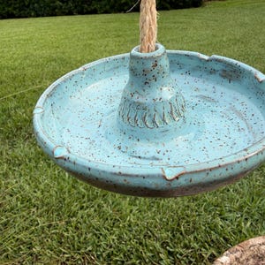 May include: A light blue ceramic hanging bird feeder with dark brown speckles. The circular dish features a central raised pillar with carved decorative grooves at the base. A thick, natural-colored twisted rope passes through the center.