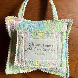 May include: A small, handmade tote bag with a quilted design in pastel colors. The bag features a handle and a central patch with the text "We love, because He first loved us." from 1 John 4:19.