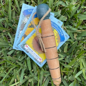 May include: A wooden seed planting tool with a blue painted top and a twine handle. The tool is sitting on top of three seed packets for Mammoth Russian Sunflower seeds. The seed packets are blue and white with a sunflower graphic and the text "American Seed Finest Quality Since 1897".