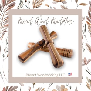 May include: Two wooden muddlers, one brown and one light brown, with a striped pattern, are crossed over each other. The text "Mixed Wood Muddlers" is above the muddlers and the text "Brandt Woodworking LLC" is below the muddlers. A small American flag is to the right of the text.