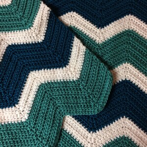 May include: A handmade crochet blanket with a chevron pattern. The blanket features alternating stripes of teal, white, and navy blue. The texture of the crochet stitches is visible, creating a cosy and textured appearance. The blanket is folded, showcasing the pattern.
