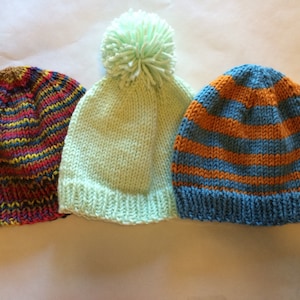 Puede incluir: Tres gorros de punto. El primero presenta un patrón de rayas multicolores en rojo, amarillo, azul y morado. El segundo es verde claro con un pompón a juego. El tercero tiene rayas horizontales naranjas y azules.
