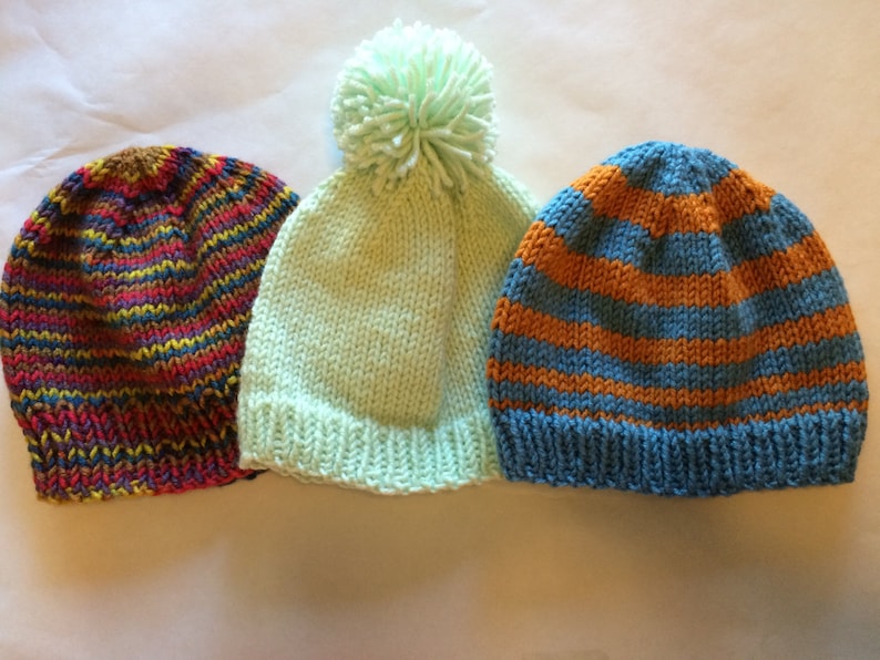 May include: Three knitted beanies in a row. The first beanie has a multicolored striped pattern in red, yellow, blue, and purple. The second is light green with a matching pom-pom. The third has horizontal orange and blue stripes.