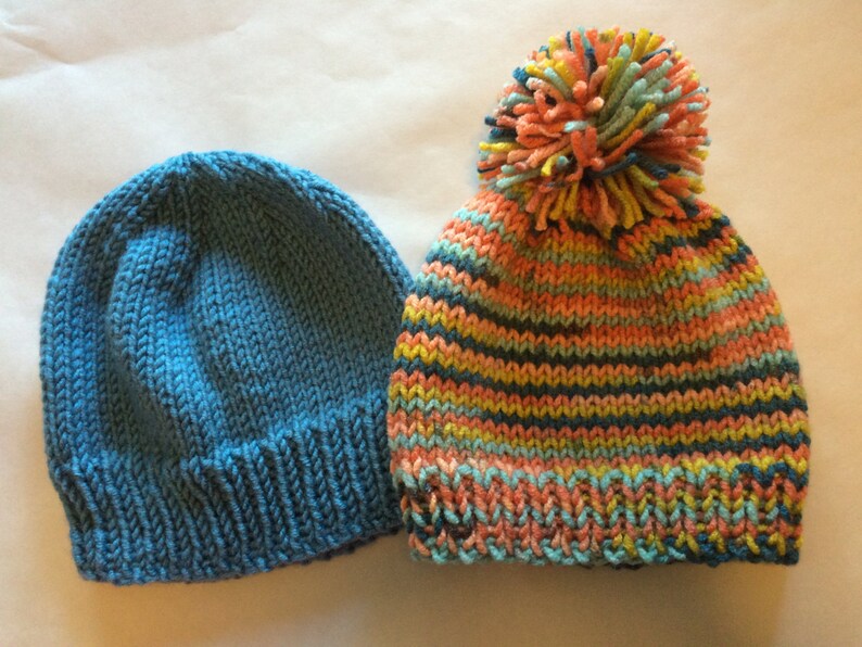 May include: Two knitted hats. One is solid blue, the other is striped with orange, teal, yellow, and navy blue. The striped hat has a large pom-pom on top made of matching yarn colors. Both hats are made of soft yarn.