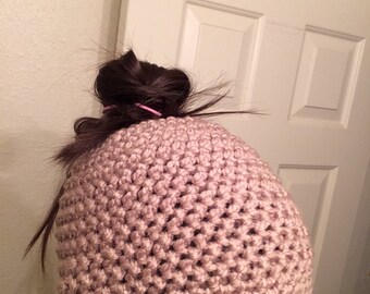 May include: A light brown, handmade crochet beanie hat. The hat is made with a textured stitch pattern and is worn on the head. The hat is a neutral color and appears to be warm and comfortable.