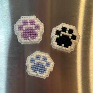 May include: Three handmade paw print magnets. One magnet features a purple paw print, another has a black paw print, and the third has a blue paw print. Each paw print is surrounded by a white border.