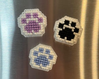 May include: Three handmade paw print magnets. One magnet features a purple paw print, another has a black paw print, and the third has a blue paw print. Each paw print is surrounded by a white border.